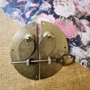 May include: A pair of antique brass cabinet door latches with a keyhole and a heart-shaped key. The latches are round and have a decorative design.