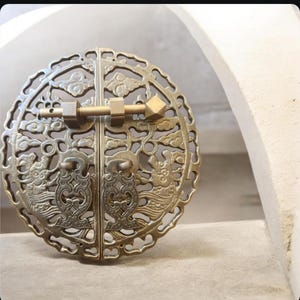 May include: A circular brass cabinet pull featuring an intricate openwork design of stylized clouds and mythical creatures. The hardware consists of two symmetrical halves joined by a horizontal sliding pin mechanism with a diamond-shaped end.