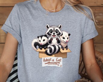 May include: Gray t-shirt with a graphic of three cute animals in a cardboard box. The animals are a raccoon, a possum, and another possum. The text on the box says "Adopt a Cat".