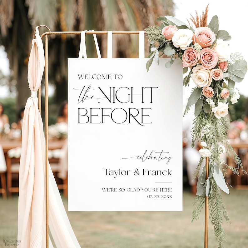 May include: A wedding sign with the words "WELCOME TO the NIGHT BEFORE" in black on a white background. The sign is adorned with a floral arrangement of pink and white roses and foliage. The names "Taylor & Franck" and the date are also included.
