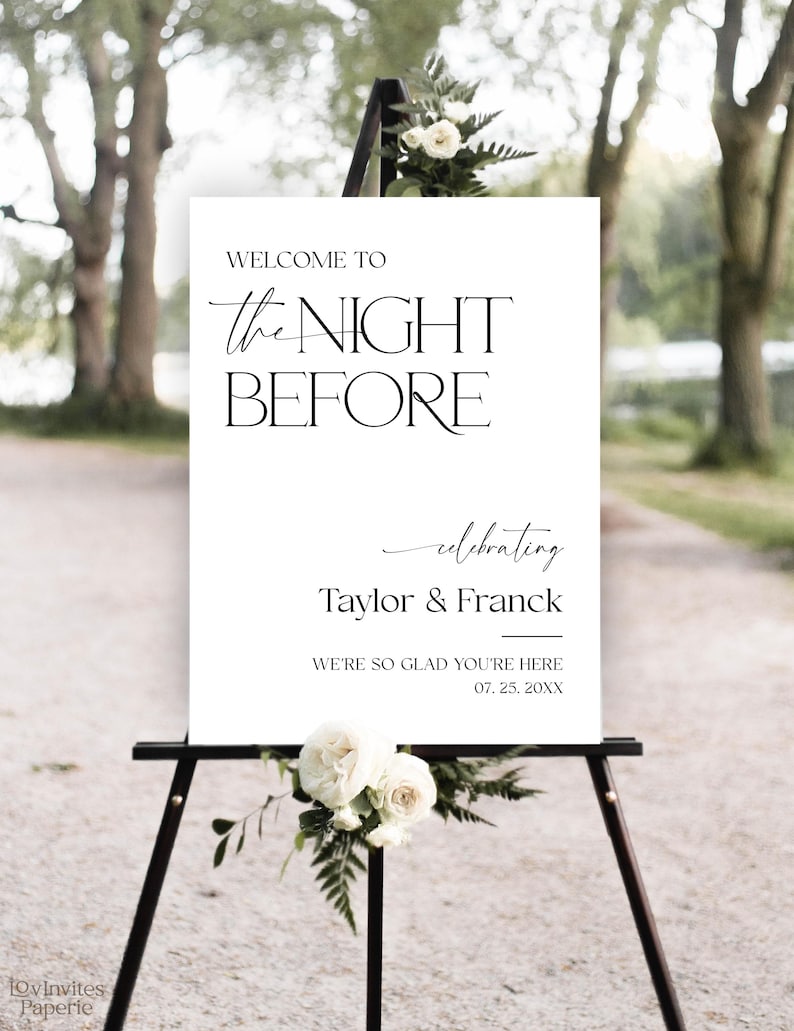 May include: A white sign on a black easel with the text "WELCOME TO the NIGHT BEFORE celebrating Taylor & Franck". The sign is decorated with white roses and greenery, suitable for a wedding or event.