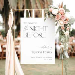 May include: A wedding welcome sign with the text "WELCOME TO the NIGHT BEFORE" in black. The sign is decorated with a floral arrangement of pink and white roses and greenery. The sign also includes the names "Taylor & Franck" and the date.