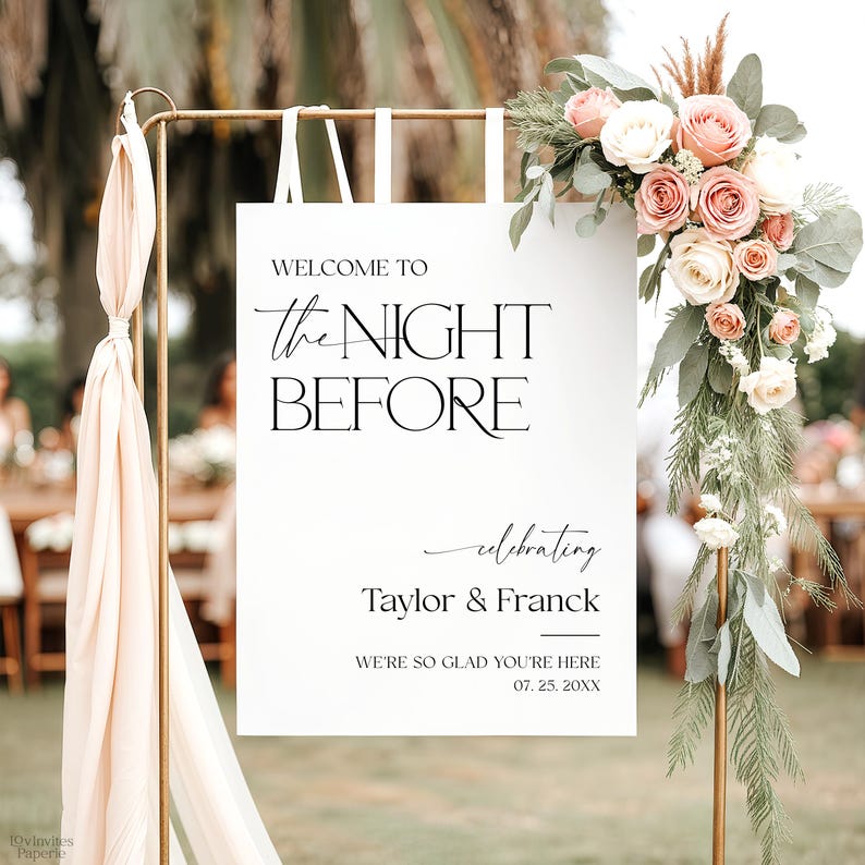 May include: A wedding welcome sign with the text "WELCOME TO the NIGHT BEFORE" in black. The sign is decorated with a floral arrangement of pink and white roses and greenery. The sign also includes the names "Taylor & Franck" and the date.
