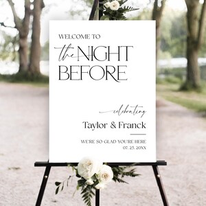 May include: A white sign on a black easel with the text "WELCOME TO the NIGHT BEFORE celebrating Taylor & Franck". The sign is decorated with white roses and greenery, suitable for a wedding or event.