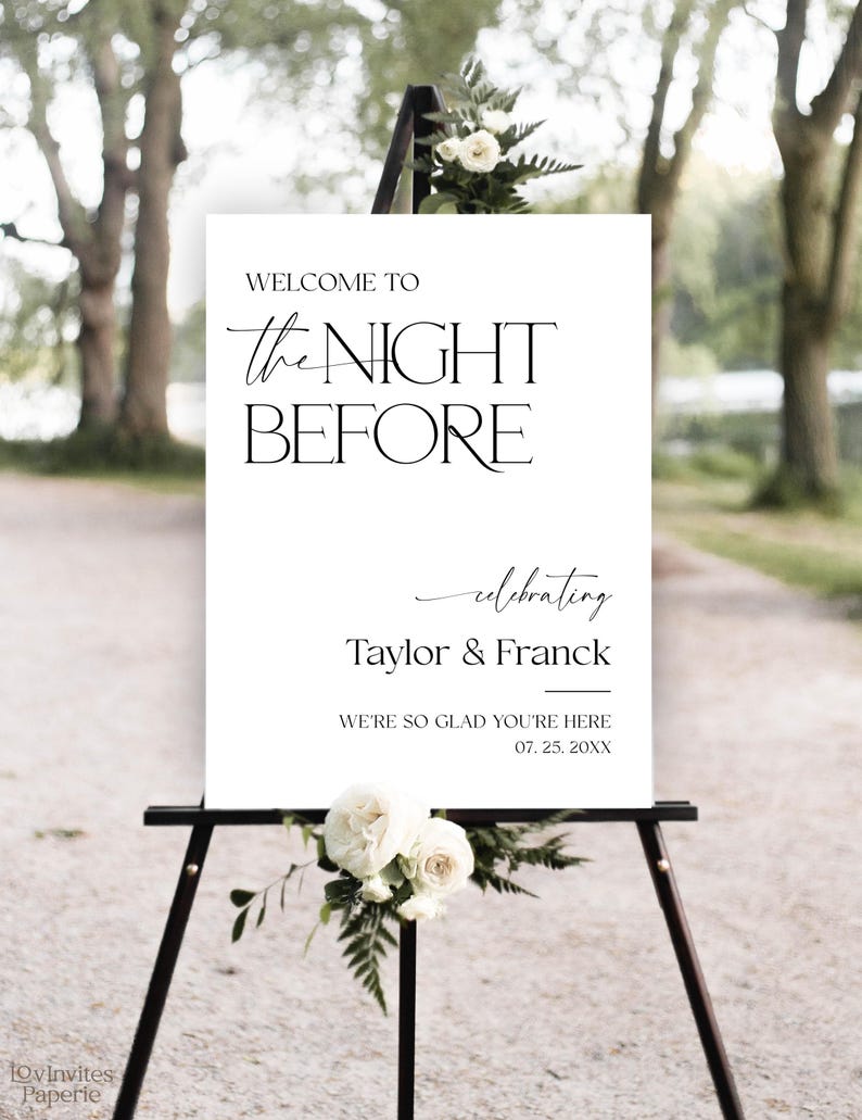 May include: A white sign on a black easel with the text "WELCOME TO the NIGHT BEFORE celebrating Taylor & Franck". The sign is decorated with white roses and greenery, suitable for a wedding or event.