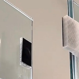 May include: A close-up of a glass shower door with a clear, rectangular hinge and a white, textured bumper. The door is attached to a metal pole. The background is a light beige wall.