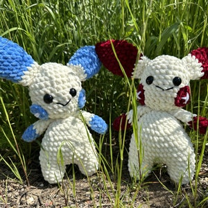 Könnte beinhalten: Zwei gehäkelte Plüschtiere, die Eevee, einem Pokémon-Charakter, ähneln. Eines ist weiß mit blauen Ohren und das andere ist weiß mit roten Ohren. Beide stehen in einem grasbewachsenen Bereich.