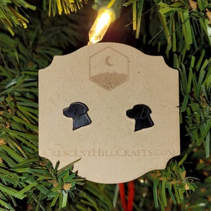 May include: Black dog head stud earrings on a tan card with a hexagonal design and the text CRESCENTHILLCRAFTS.COM. The earrings are displayed on a Christmas tree with a string of lights.