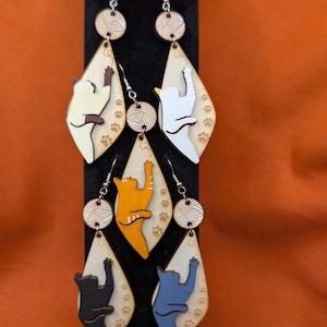 May include: A set of six cat-themed dangle earrings. Each earring features a wooden, teardrop-shaped pendant with a cat silhouette climbing towards a ball of yarn. The cats are in various colors, including white, orange, black, and blue. Paw prints are etched on the pendants.