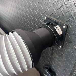 May include: A black and white corrugated hose is connected to a black electrical outlet on a textured, dark gray metal surface. The hose is attached to a black adapter.