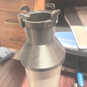 May include: A black plastic vacuum cleaner hose adapter with a flared base and a ridged, cylindrical body. The top features four curved tabs. The adapter is designed to connect to a vacuum cleaner hose.