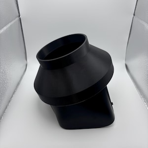 May include: A black, angled, plastic object with a flared top and a rectangular base. The object is positioned diagonally against a white background, with a studio light visible in the upper left corner.