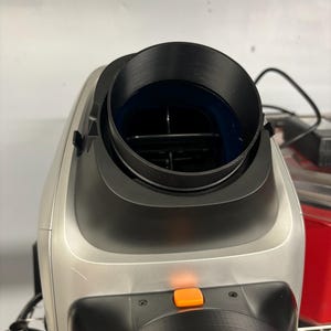 May include: A silver and black appliance with a cylindrical black opening at the top. An orange button is visible on the front. The appliance has a modern design and appears to be electronic.