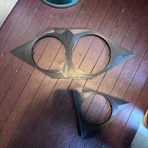 May include: Three-dimensional, dark gray geometric shapes. One large piece has two circular openings and a pointed, diamond-like shape. A smaller, triangular piece with a circular opening is also present. The objects are on a wooden surface.