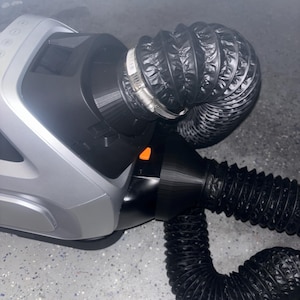 May include: A silver and black air purifier with a black flexible hose attached to the side. The hose is connected to the air purifier with a silver clamp.