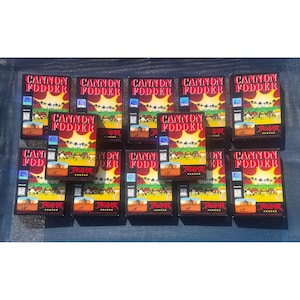 May include: A collection of boxed video games, featuring the title "CANNON FODDER" in red and yellow text. The boxes have a landscape graphic with soldiers and a desert scene. The game is for the Jaguar console.