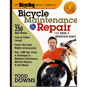 May include: The Bicycling Guide to Complete Bicycle Maintenance & Repair for Road & Mountain Bikes, 5th Edition by Todd Downs. The book cover features a person in an orange shirt working on a tan mountain bike with black tires and handlebars.