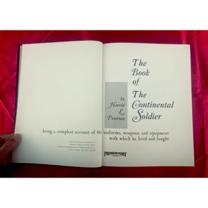 May include: An open book with the title "The Book of The Continental Soldier" by Harold L. Peterson. The book's pages are off-white, with black text. The book is about uniforms, weapons, and equipment.