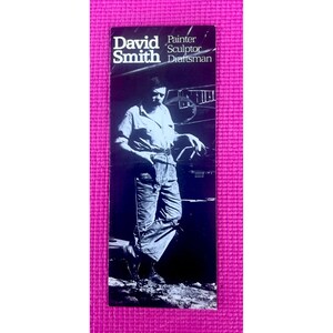 May include: Black and white promotional card featuring David Smith, a painter, sculptor, and draftsman. The card displays a portrait of Smith in work attire, leaning against a dark object. The text "David Smith" is prominently displayed at the top.