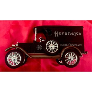 May include: A dark brown vintage Hershey's Milk Chocolate delivery truck toy with silver wheels and accents. The truck has the Hershey's logo and "Milk Chocolate" text on the side. The toy is set against a red background.