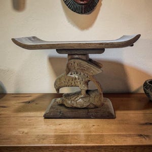 May include: A wooden decorative stand with a carved eagle base and a rectangular top. The stand is made of light brown wood and is displayed on a wooden surface. Other carved wooden objects and a mask are visible in the background.