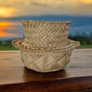 May include: Two woven straw hats, one stacked on top of the other, sit on a wooden surface. The hats have a natural tan color and a textured, basket-weave design. The background features a blurred sunset over a green field.