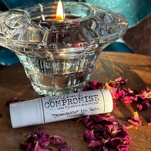 Puede incluir: Tubo blanco de bálsamo labial "Dependable" con las palabras "Without Compromise" y "Low Tox Bath & Beauty Care" en la etiqueta. El tubo está sobre una superficie de madera con pétalos de rosa secos.