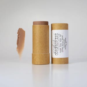 May include: A brown cardboard tube with a white label that reads "Without Compromise Low Tox Bath & Beauty Care "Just Glow With It" Tinted Tallow Facial Moisturizer Golden Goddess 50 ml". The tube is next to a brown cardboard tube with a brown balm inside. A small amount of the balm is on a white surface.