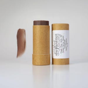 May include: A brown cardboard tube with a white label that reads "Without Compromise Low Tox Bath & Beauty Care "Just Glow With It" Tinted Tallow Facial Moisturizer Ebony Elegance". The tube contains a brown tinted balm.