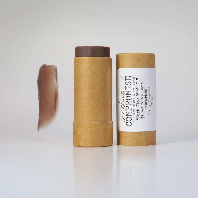 May include: A brown cardboard tube with a white label that reads "Without Compromise Low Tox Bath & Beauty Care "Just Glow With It" Tinted Tallow Facial Moisturizer Ebony Elegance". The tube contains a brown tinted balm.