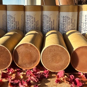 May include: Close-up of ten brown cardboard tubes with white text that reads "PROMISE KATH & BEAUTYCARE Glow With It" and "Glow Mith It" on the tubes. The tubes are filled with a brown product and are sitting on a wooden surface with red flower petals scattered around them.