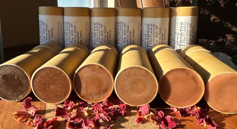 May include: Close-up of ten brown cardboard tubes with white text that reads "PROMISE KATH & BEAUTYCARE Glow With It" and "Glow Mith It" on the tubes. The tubes are filled with a brown product and are sitting on a wooden surface with red flower petals scattered around them.