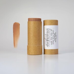 May include: A brown cardboard tube with a white label that reads "Without Compromise Low Tox Bath & Beauty Care "Just Glow With It" Tinted Tallow Facial Moisturizer Bronze Beauty 50 mL". The tube contains a brown tinted balm.