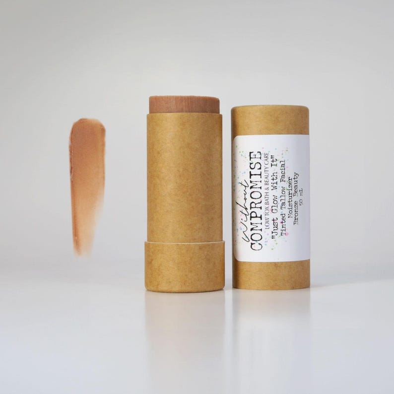 May include: A brown cardboard tube with a white label that reads "Without Compromise Low Tox Bath & Beauty Care "Just Glow With It" Tinted Tallow Facial Moisturizer Bronze Beauty 50 mL". The tube contains a brown tinted balm.