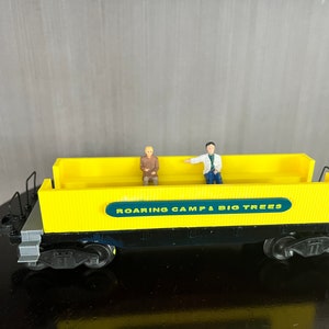 May include: Yellow toy train car with black wheels and a green sign that reads "Roaring Camp & Big Trees". Two small figures are sitting on the car.