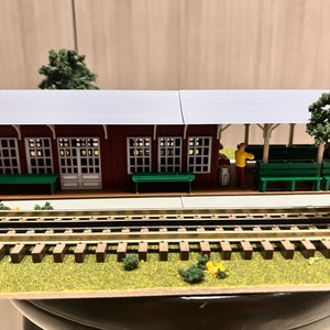 May include: A model train station with a gray roof, red walls, and green benches. The station has a small figure of a person standing near a trash can. The station is set up on a model train track with brown wooden ties and gold metal rails.