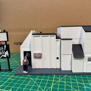 以下が含まれることがあります： 「Bada Bing」の看板が付いた白い建物のミニチュアモデル。建物は白と黒の屋根とトリム。小さな男性のフィギュアが前に立っています。空白のスペースのある看板も存在します。モデルは緑色の表面にあります。