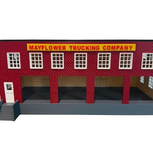 May include: A model building featuring red brick walls, a yellow sign reading MAYFLOWER TRUCKING COMPANY, white window frames, and a white door with stairs. The structure has an open ground level with four bays and a gray base foundation.