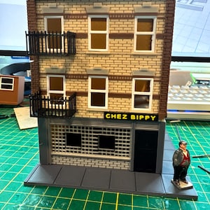 May include: A miniature brick building with a black awning and the words "CHEZ BIPPY" in yellow letters. The building has two balconies with black railings and windows with white frames. A small figure of a person in a brown jacket and black pants stands on the sidewalk in front of the building.