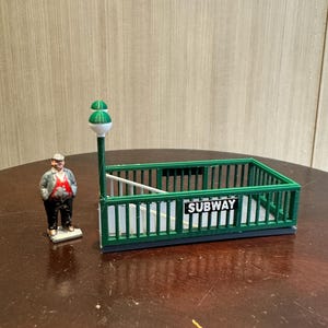 以下が含まれることがあります： 白い文字で「SUBWAY」と書かれたミニチュアの緑色の地下鉄の入り口。帽子と緑と赤のベストを着た男性の姿が近くに立っています。緑と白の街灯も存在します。