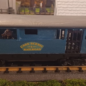 Pode incluir: Um vagão de trem modelo azul com telhado preto e uma placa que diz "Cass Scenic Railroad". O vagão tem janelas e uma porta no lado. Há pequenas figuras de pessoas dentro do vagão.