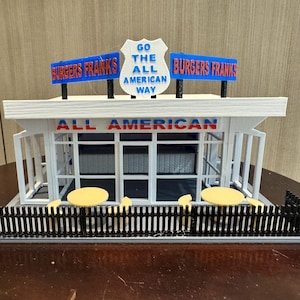 Puede incluir: Un modelo blanco de un restaurante con un letrero rojo y azul que dice "Go the All American Way" y "Burgers Franks" a cada lado. El restaurante tiene un techo blanco y una valla de madera negra. Hay dos mesas amarillas con sillas fuera del restaurante.