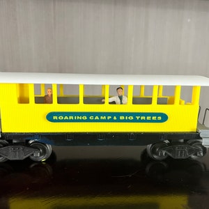 May include: Yellow toy train car with a white roof and black wheels. The car has windows and a sign that reads "ROARING CAMP & BIG TREES".