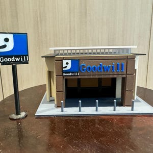 Pode incluir: Um modelo em miniatura de uma loja Goodwill com uma placa azul e branca que diz "Goodwill". O modelo é feito de papelão marrom e branco e tem um telhado branco. Há seis pequenos postes cinzentos na frente da loja.