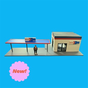 May include: A miniature model train station with a white building, blue and red accents, and the word "Amtrak" on the side. A small figure of a person stands under a blue and red awning.