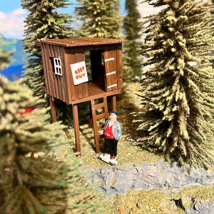 May include: A miniature wooden treehouse with a ladder and a sign that reads "Keep Out". A miniature person stands in front of the treehouse.