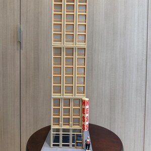 May include: A miniature model of a tall building with multiple windows and a sign that reads "HESS" in red letters. A small figure stands near the base of the building. The building is beige with brown window frames.