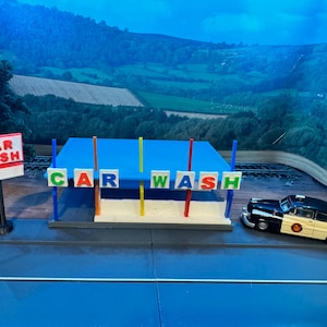 May include: A miniature car wash set with a blue roof and a white base. The sign reads "CAR WASH" in red letters. A white car with a black roof and a black and white police-style design is driving into the car wash.
