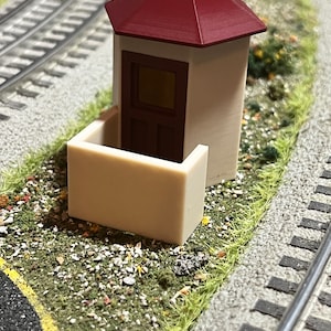 May include: A small, white, model train station with a red roof and a brown door. The station is sitting on a bed of green grass and gravel.