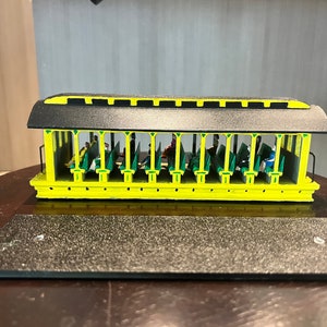 Pode incluir: Um vagão de trem modelo amarelo com telhado preto e detalhes verdes. O vagão está cheio de passageiros em miniatura e tem um interior detalhado.